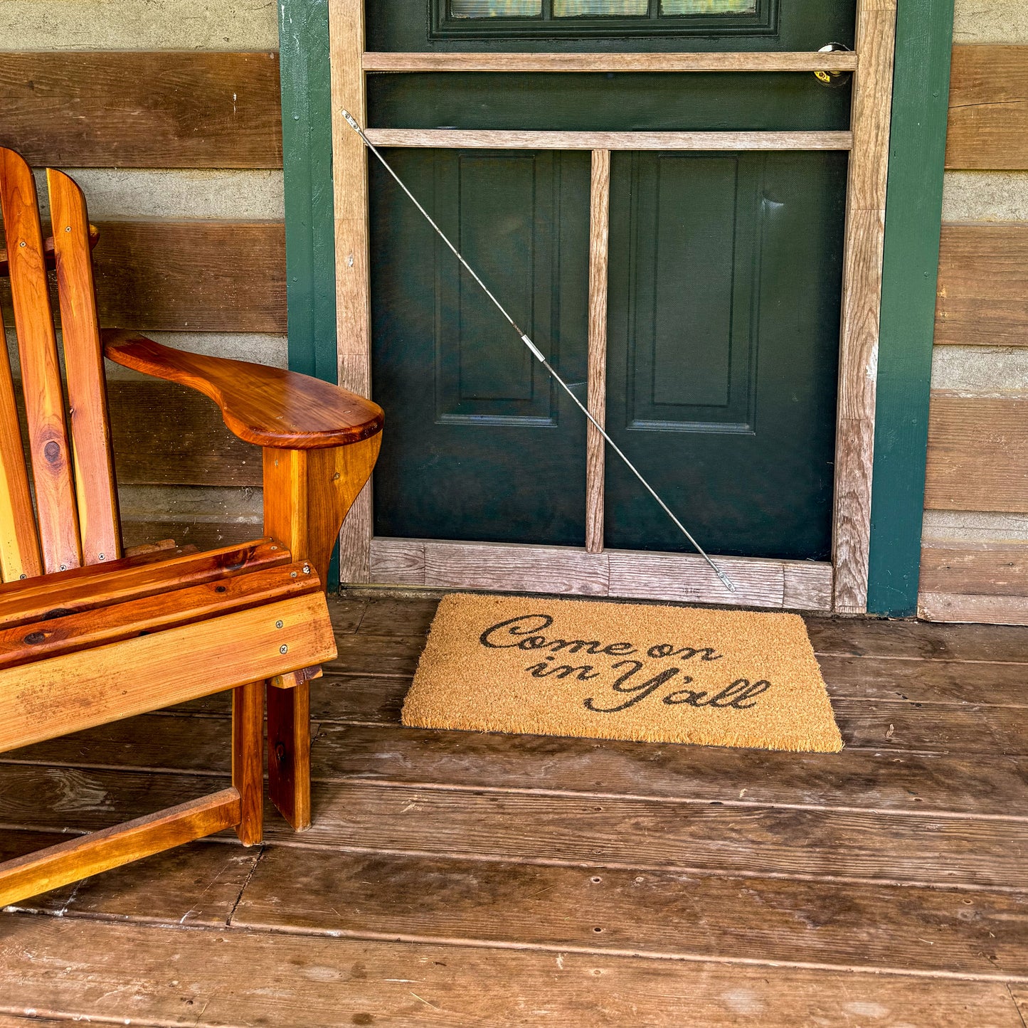 Wooden chair on a porch with a green door and 'Come on in Y'all' doormat.