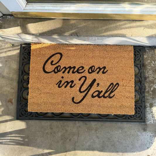 Doormat with 'Come on in Y'all' text on a concrete floor.